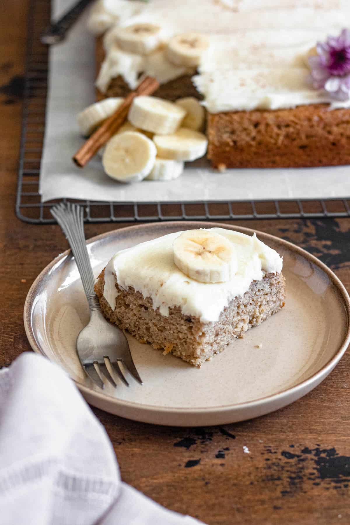 Slice of banana cake with cream cheese frosting on a plate with a banana slice garnished on top and a fork on the side. 