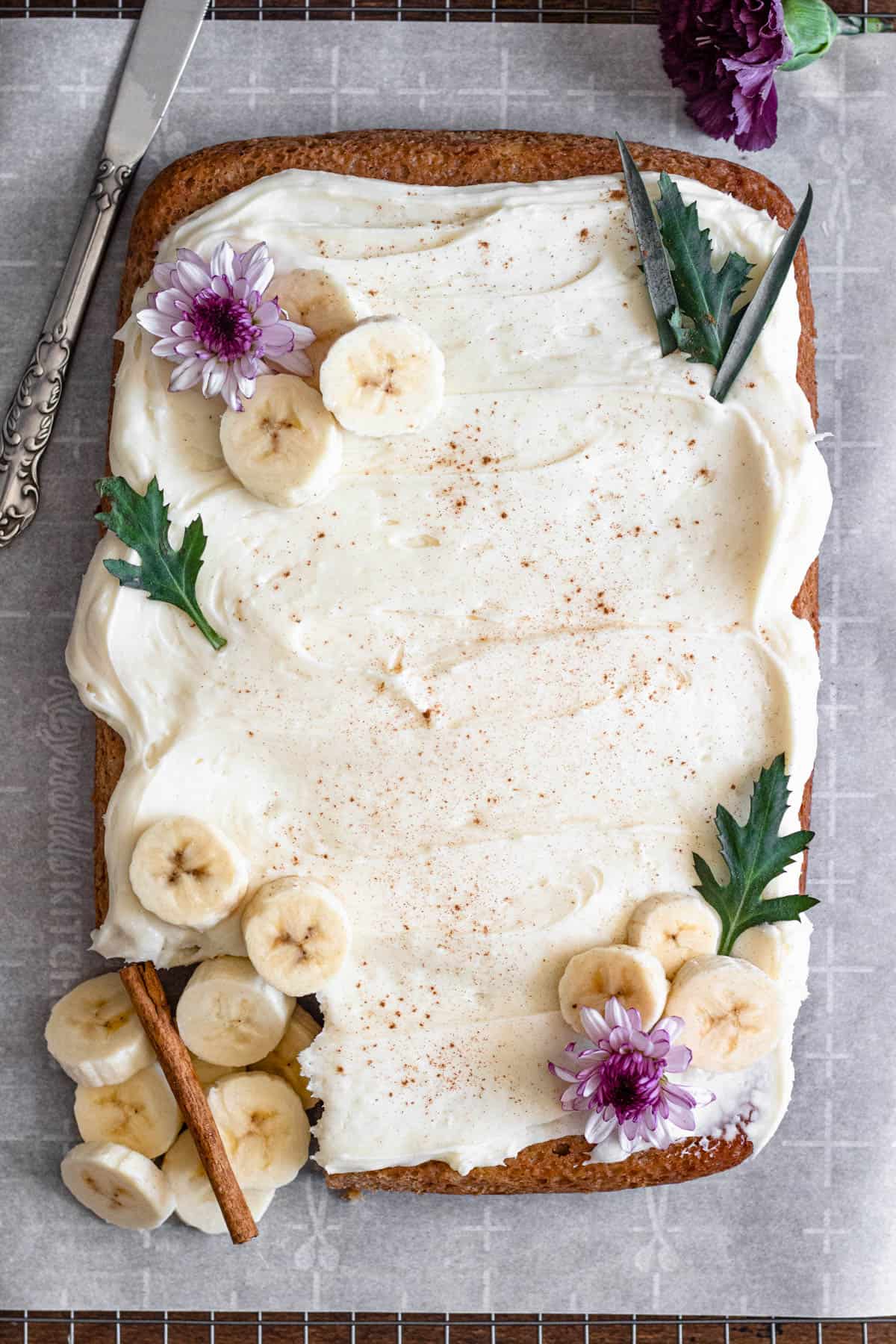Full banana cake covered in cream cheese frosting and garnished with banana slices and small purple flowers. 
