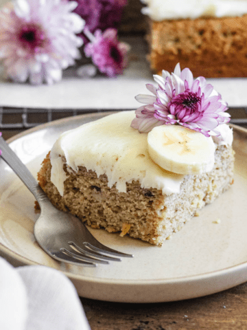 Slice of cream cheese frosted banana cake with fresh banana slices on top and a pink flower.