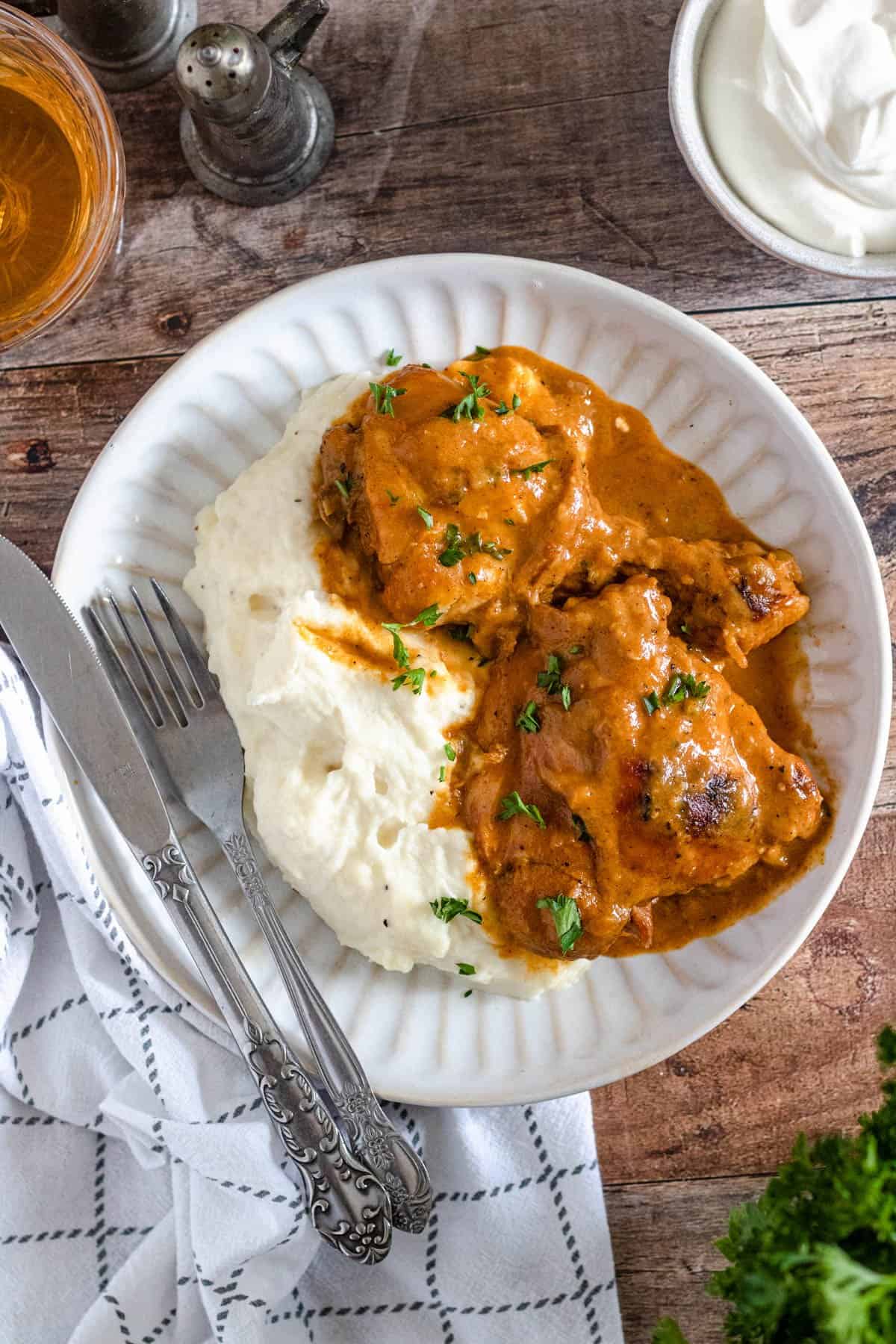 Fork and knife on a plate of chicken paprikash on a plate and a side of mashed potatoes. 