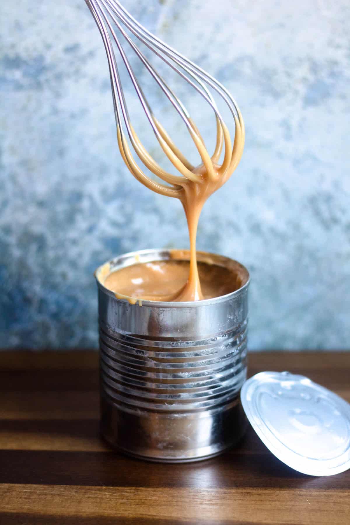 A whisk coming out of an open can of thickened caramel colored sweetened condensed milk and drizzling back into it. 