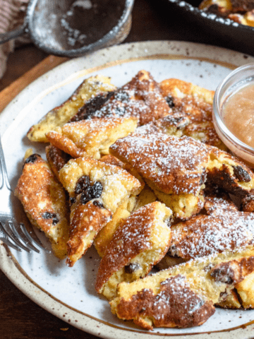 Large pieces of Kaiserschmarrn on a plate topped with powder sugar and a small bowl of applesauce alongside.