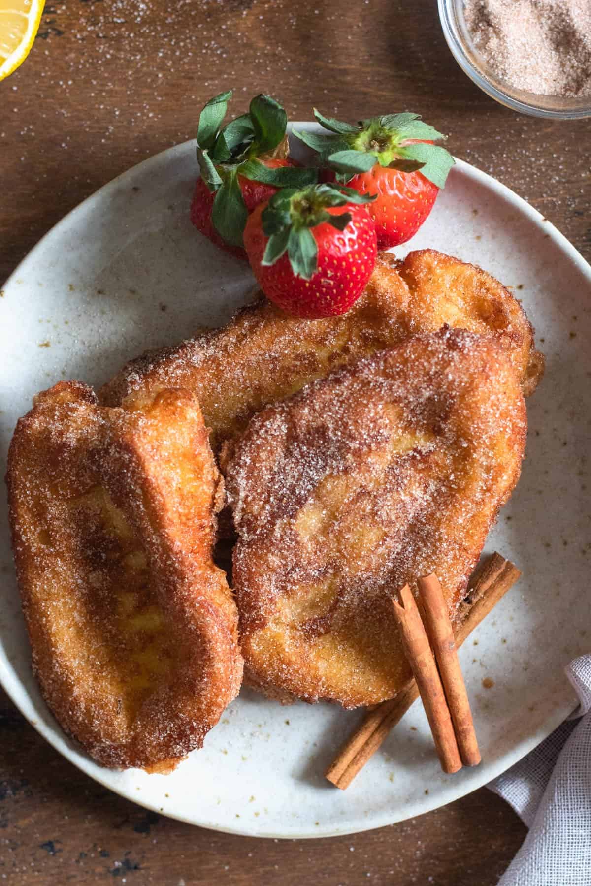 A plate with a couple Torrijas and 3 whole strawberries on the side and 2 cinnamon sticks garnishing the plate. .