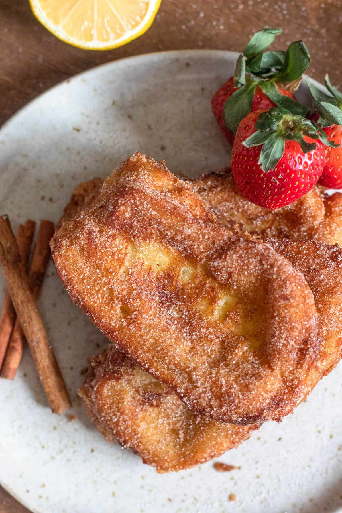 Torrijas stacked on top of one another on a plate with garnishes of strawberries and cinnamon sticks. 