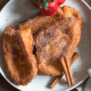 3 slices of Torrijas on a plate wtih cinnamon sticks and whole strawberries on the side.