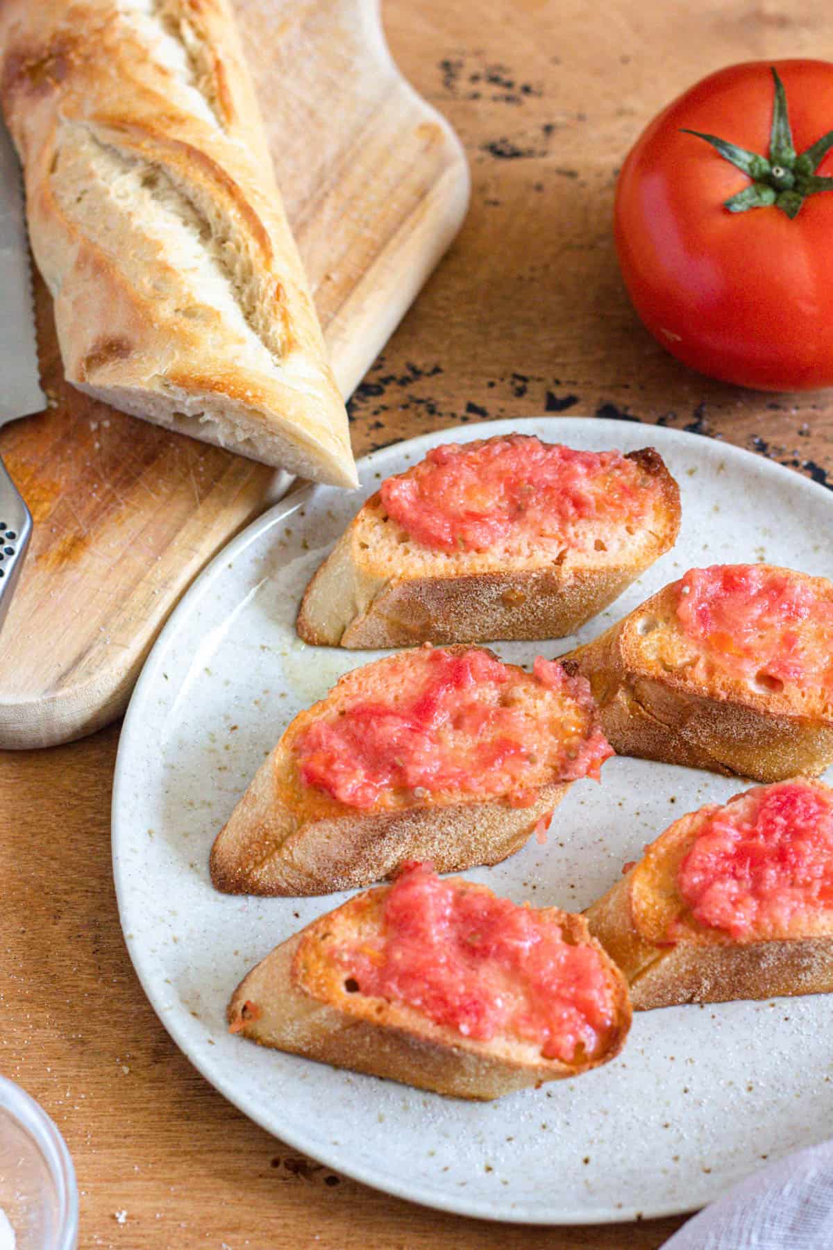 Pan con tomate served on a plate with a roma tomato and crusty bread loaf behind it on a cutting board. 