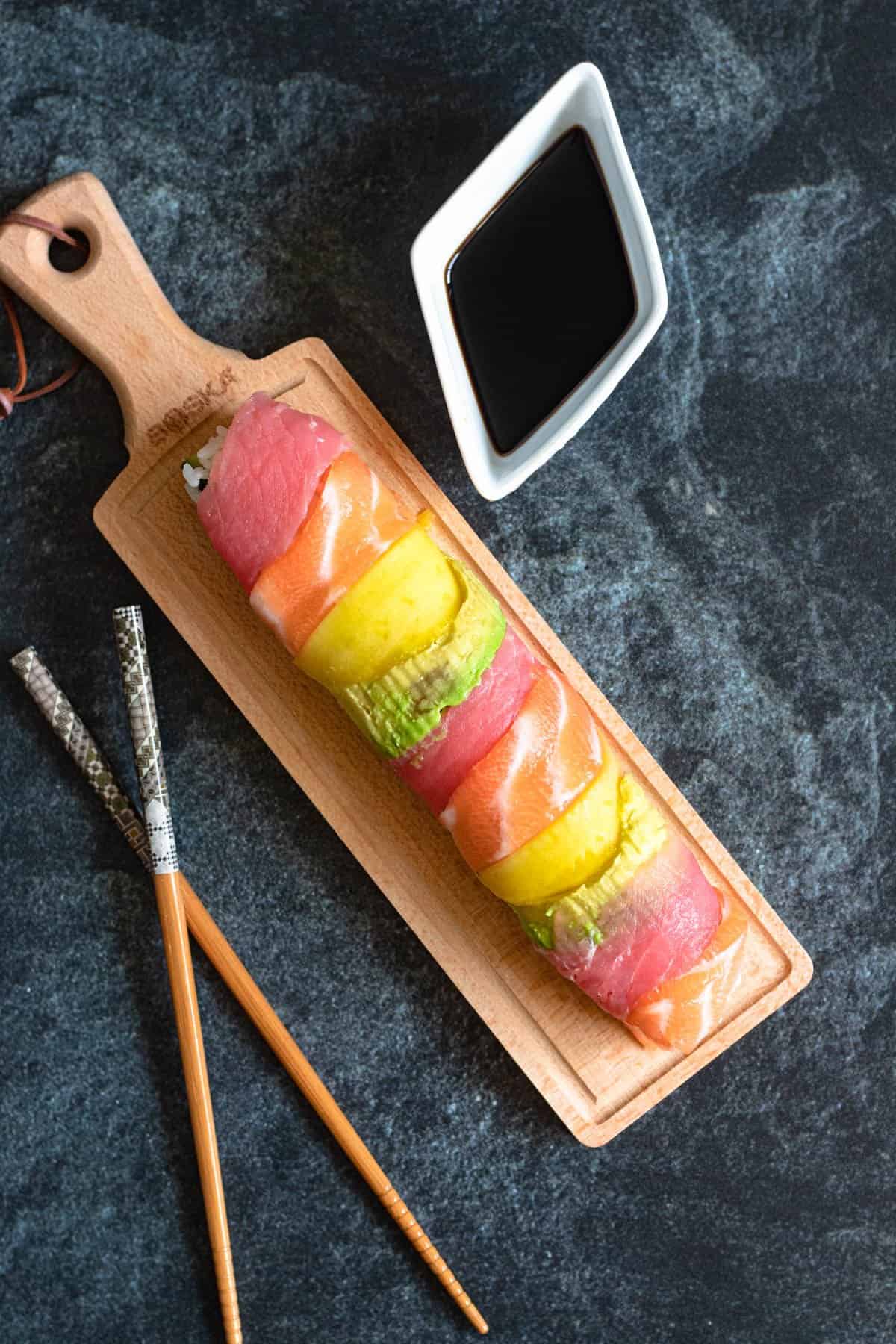 A rainbow roll displayed on a cutting board prior to cutting into slices with a side of sauce and a pair of chopsticks next to it. 
