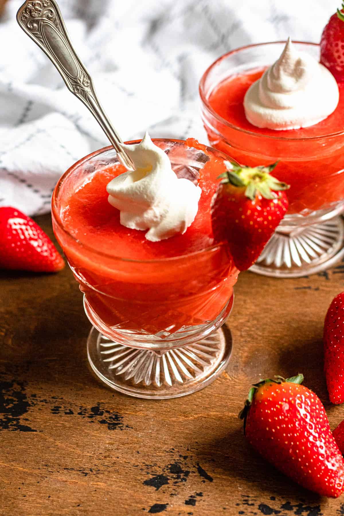 Spoons tucked into two serving glasses of strawberry kissel with a dollop of whip cream on top and a whole strawberry. 