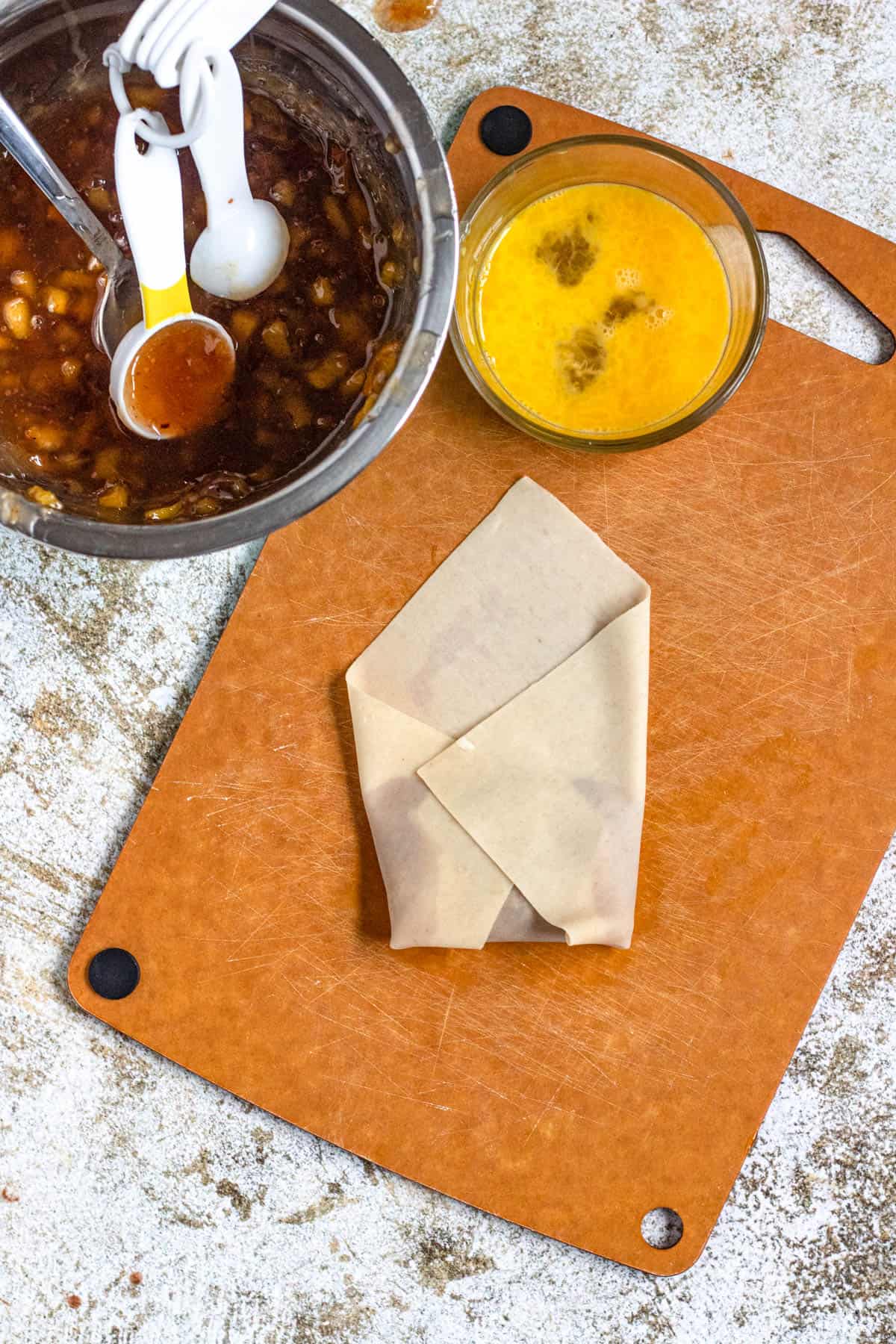An egg roll wrapper laying on a cutting board with a bit of peach cobbler filling added to it and then beginning to wrap the egg roll wrapper up so the filling is inside for peach cobbler egg rolls. 