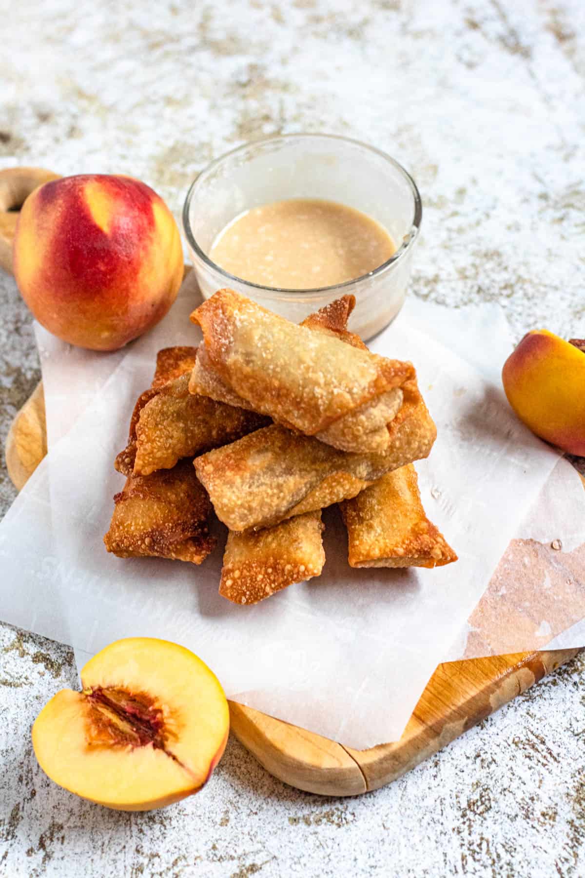 Stack of peach cobbler egg rolls sitting on a piece of parchment paper on a cutting board next to some whole peaches and vanilla dip. 