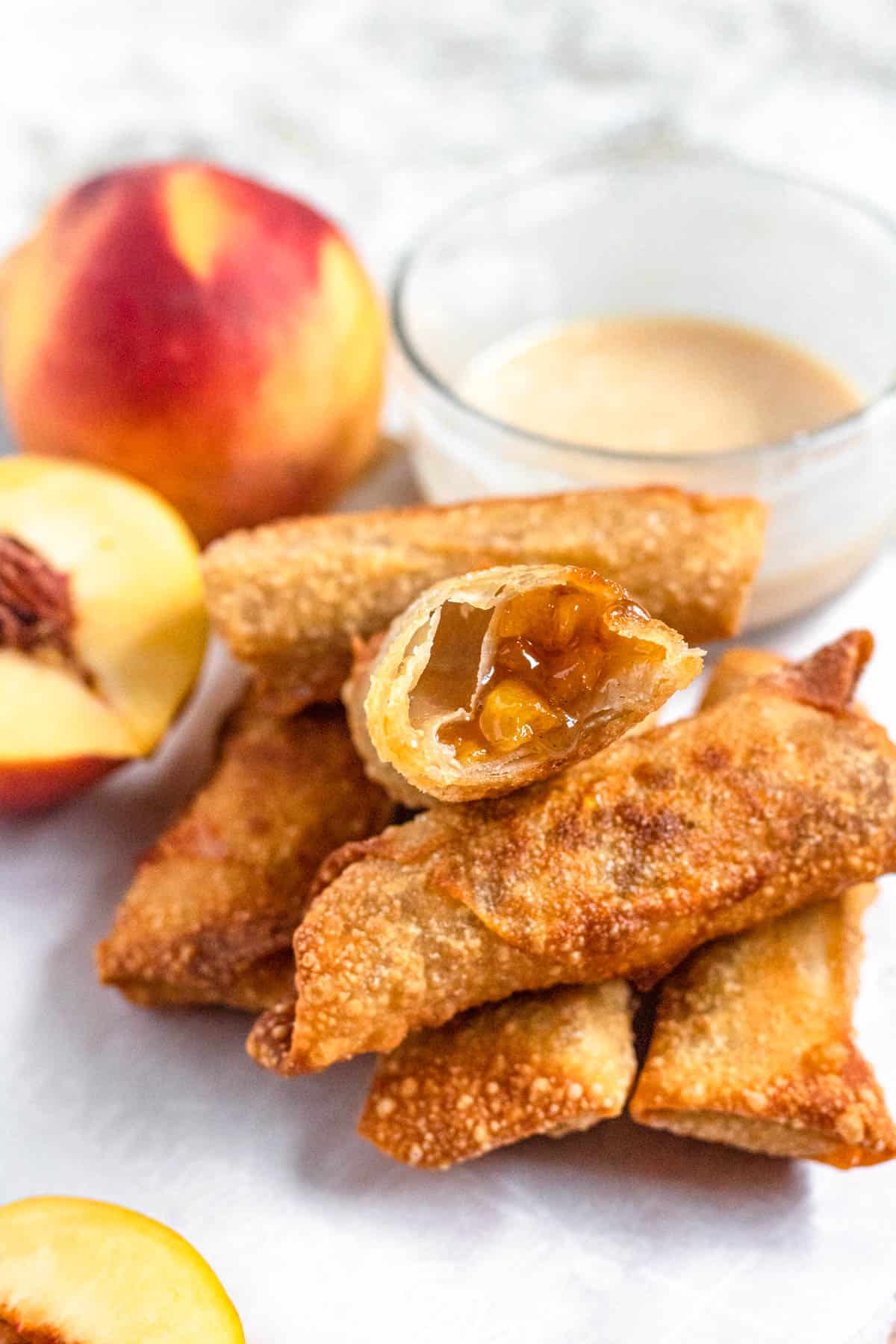 Close up of a peach cobbler egg roll with a bite taken out of it next to a halved peach and some vanilla homemade dip. 
