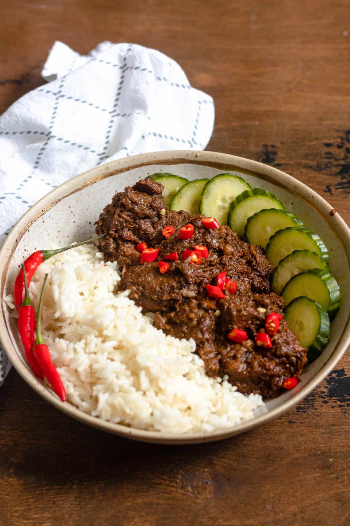 Beef rendang on a plate with sliced cucumbers and white rice and garnished with red chili pieces. 