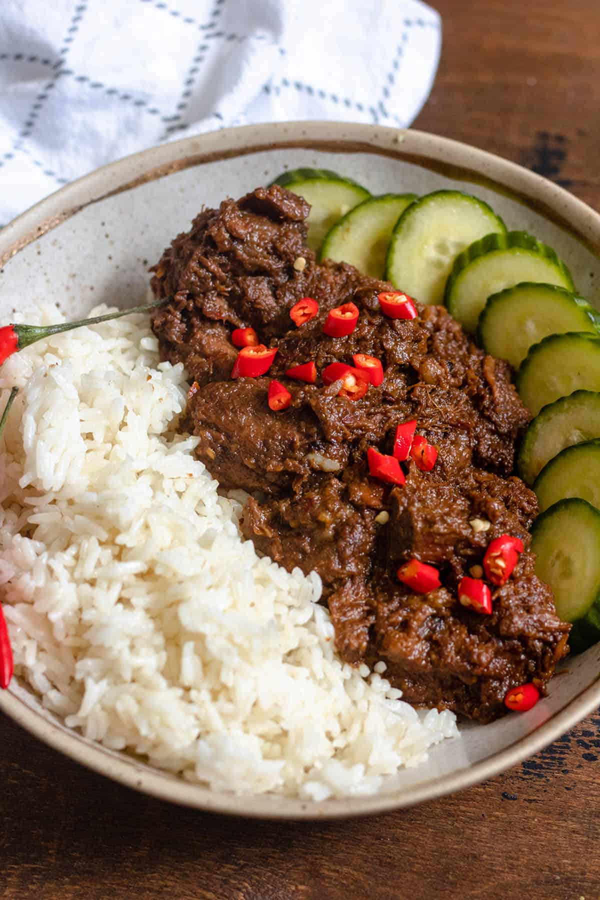 Beef rendang on a plate with sliced cucumbers and white rice and garnished with red chili pieces. 