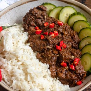Beef Rendang in a bowl with white rice, cucumbers, and chopped chilies.