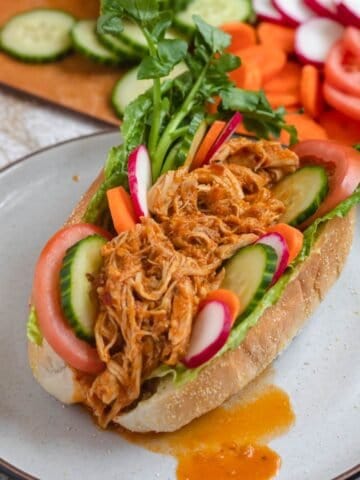 A sandwich on a plate stuffed with lettuce, tomatoes, radishes, cucumbers, carrots, and pulled chicken making a Pan con Pollo sandwich, sitting in front of a cutting board of vegetables.
