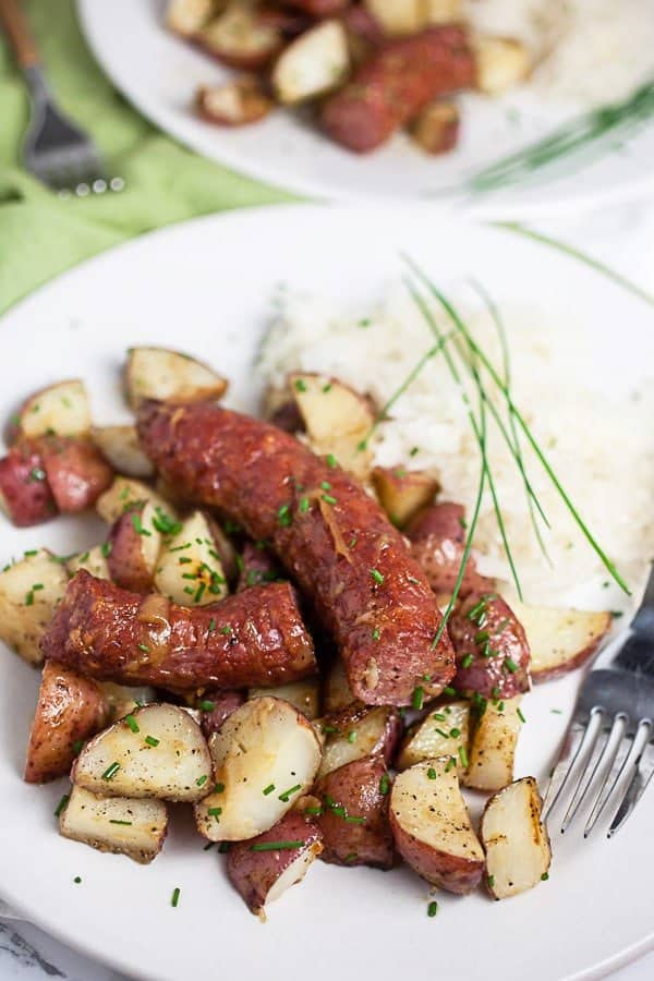 Oven roasted sausages and potatoes served on a plate with fresh herbs garnished on the side. 
