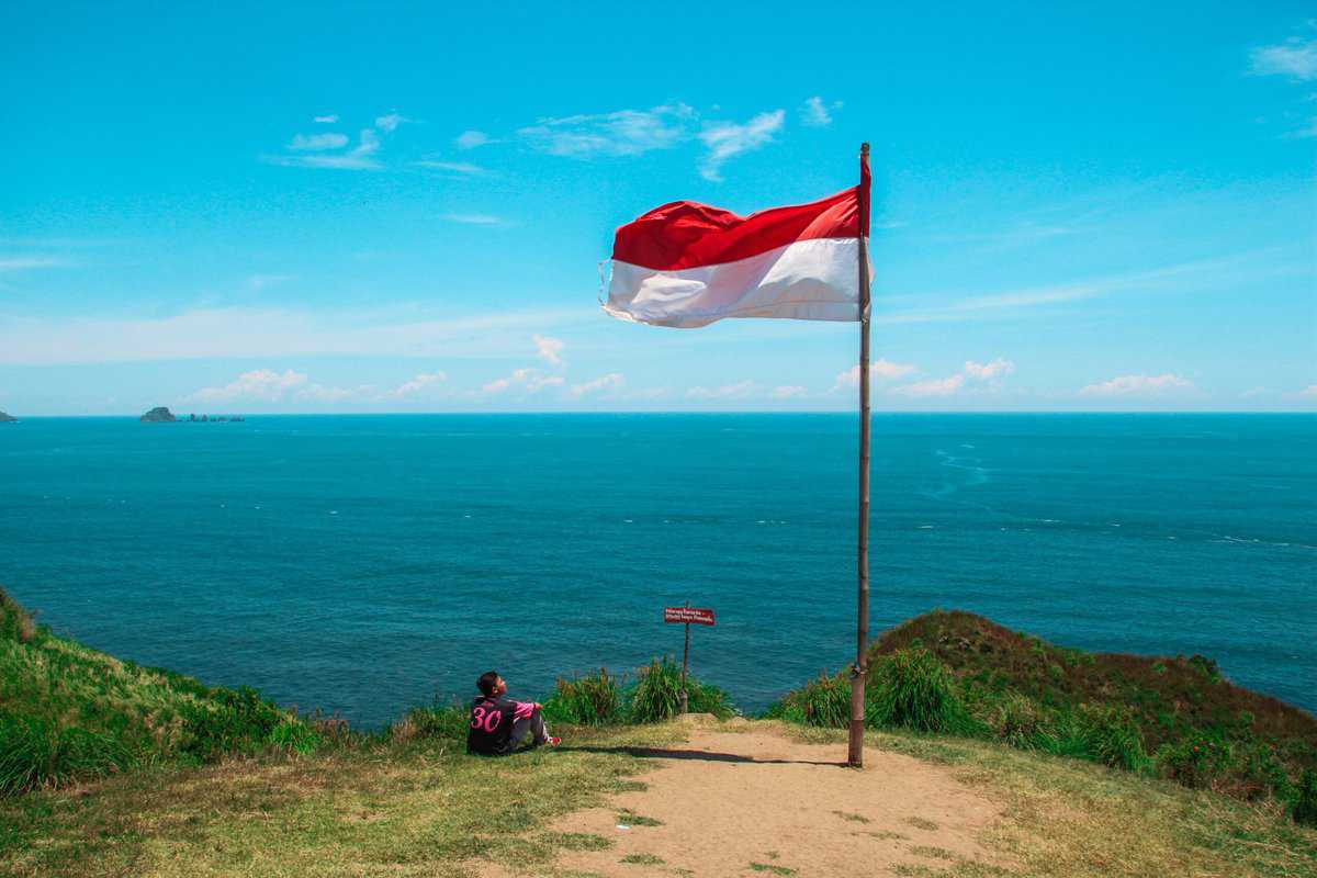 Man sitting next to an Indonesian flag by the ocean. 