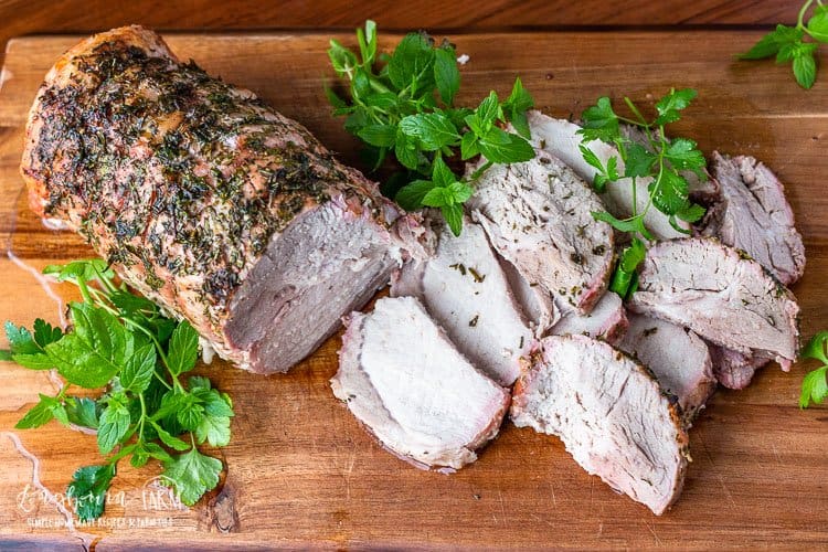 Pork loin on a cutting board with several slices cut away from it with fresh herbs around it. 