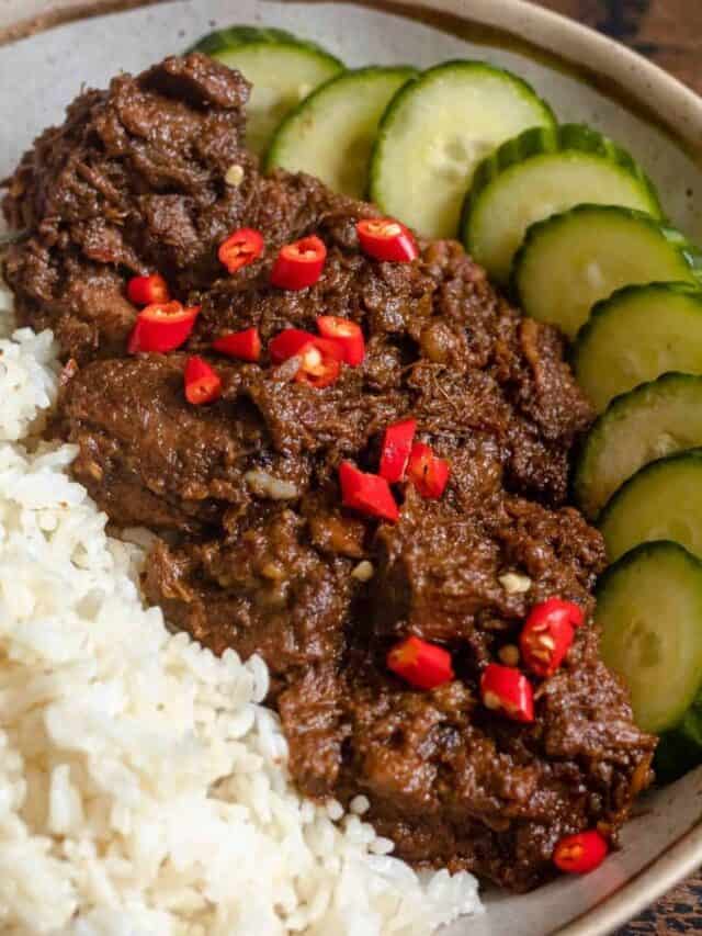 Beef rendang served on a plate with white rice and fresh cut cucumbers. 