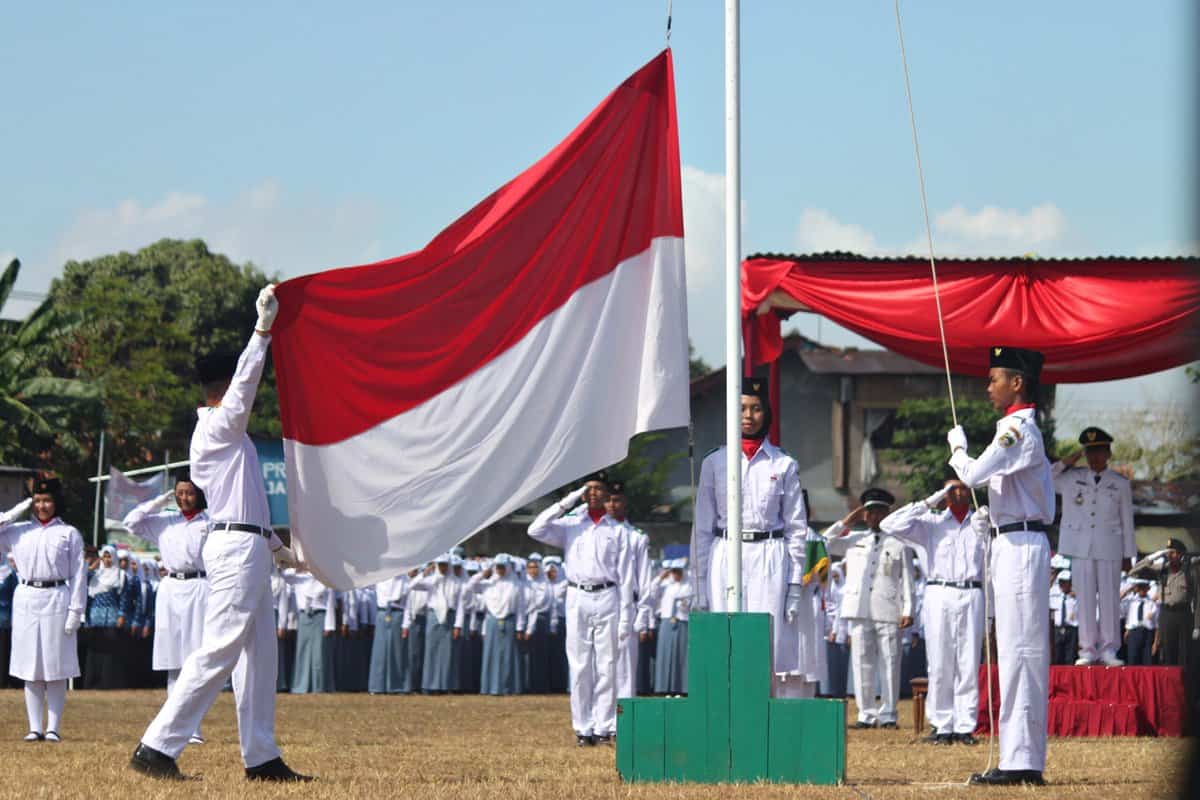 Indonesian people in uniform raising a red and white Indonesian flag while the others in uniform salute. 