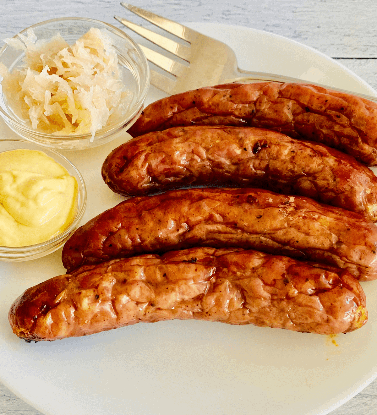 4 cooked brats on a plate next to a small bowl of dipping sauce and a small bowl of sauerkraut. 