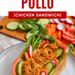 Pinterest image of Pan con Pollo on a plate, topped with lettuce and other fresh veggies, sitting in front of the cutting board with remaining toppings.