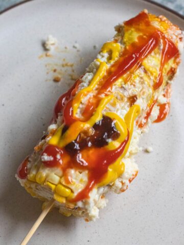 An ear of corn on a plate topped with mayonnaise, ketchup, mustard, worcestershire sauce, and cheese, making Elote Loco.