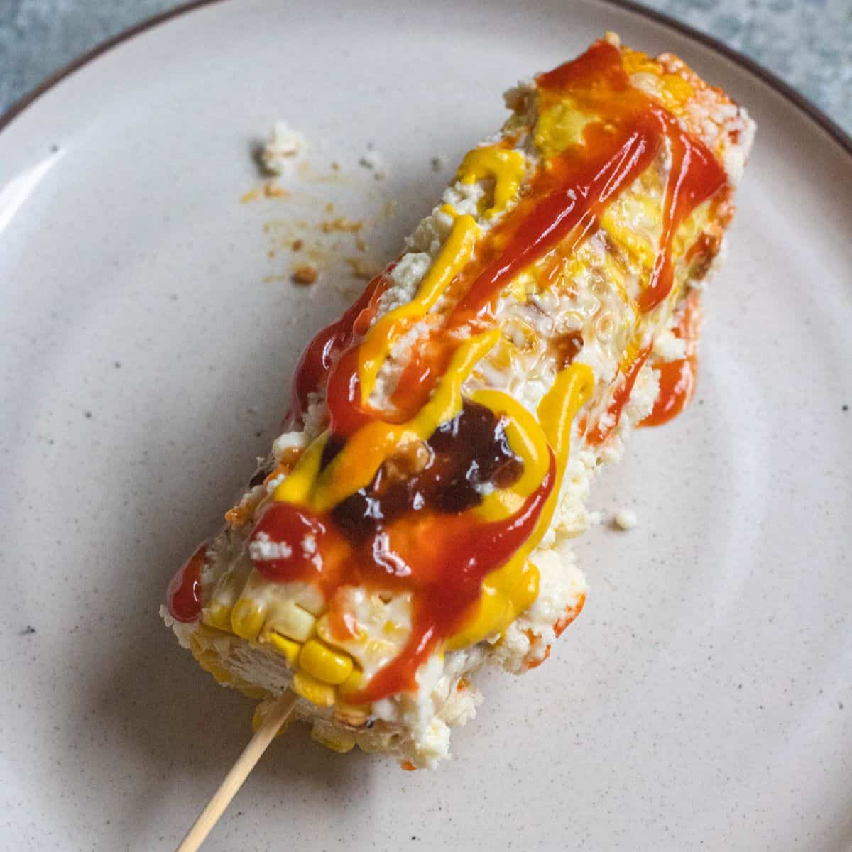 An ear of corn on a plate topped with mayonnaise, ketchup, mustard, worcestershire sauce, and cheese, making Elote Loco.