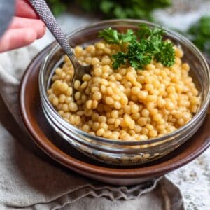A clear bowl filled with Israeli couscous and a spoon scooping it.