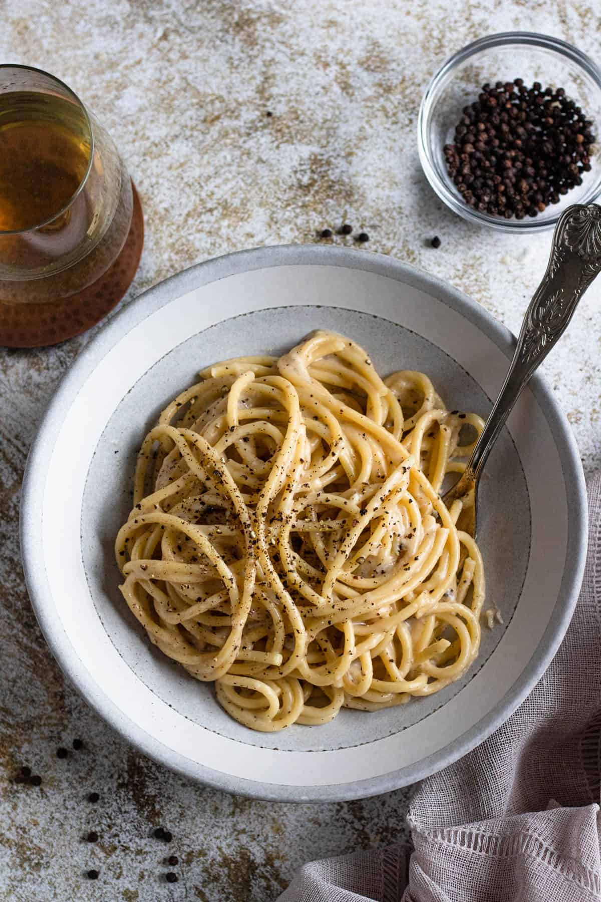 Bucatini cacio e pepe served in a bowl with a fork and a small dish of peppercorns behind it.