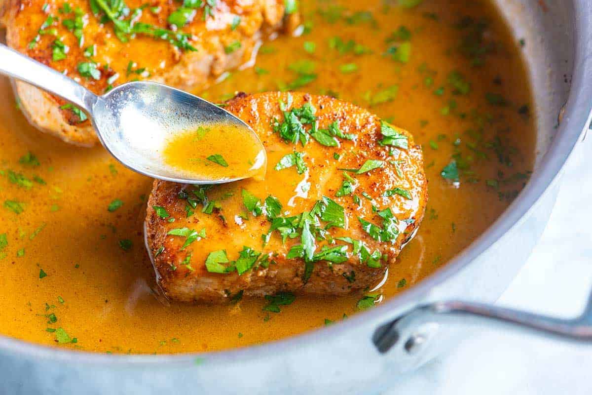 Juicy pork chops sitting in a skillet with sauce being spooned over the pork piece. 