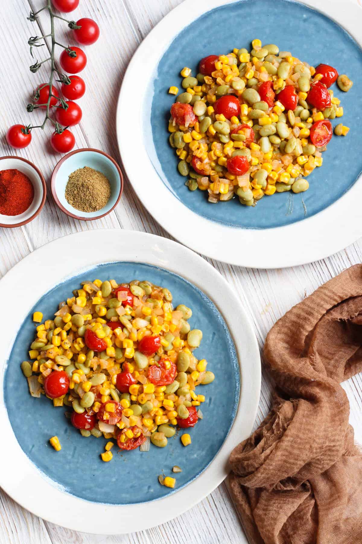 Two small blue plates with lima beans and succotash served next to cherry tomatoes and small bowls with spices.