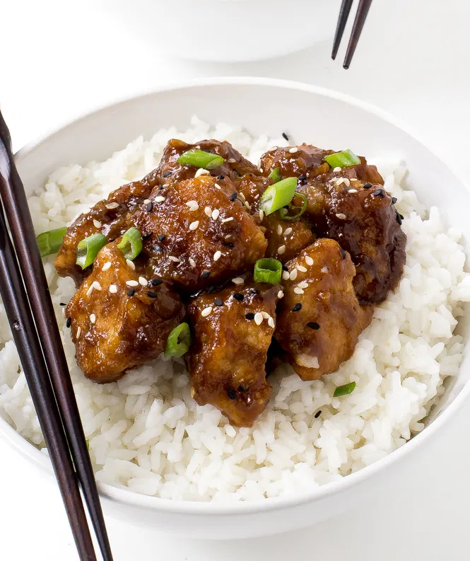 Slow cooker general tso's chicken served over a bed of white rice and a pair of chopsticks on the rim of the bowl.