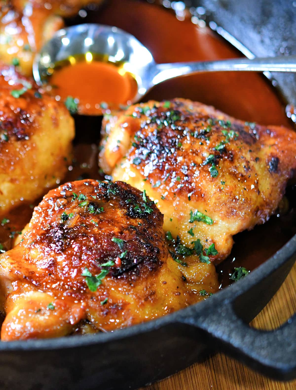 Sweet and spicy chicken thighs in a skillet with a spoon of sauce to pour over the chicken. 