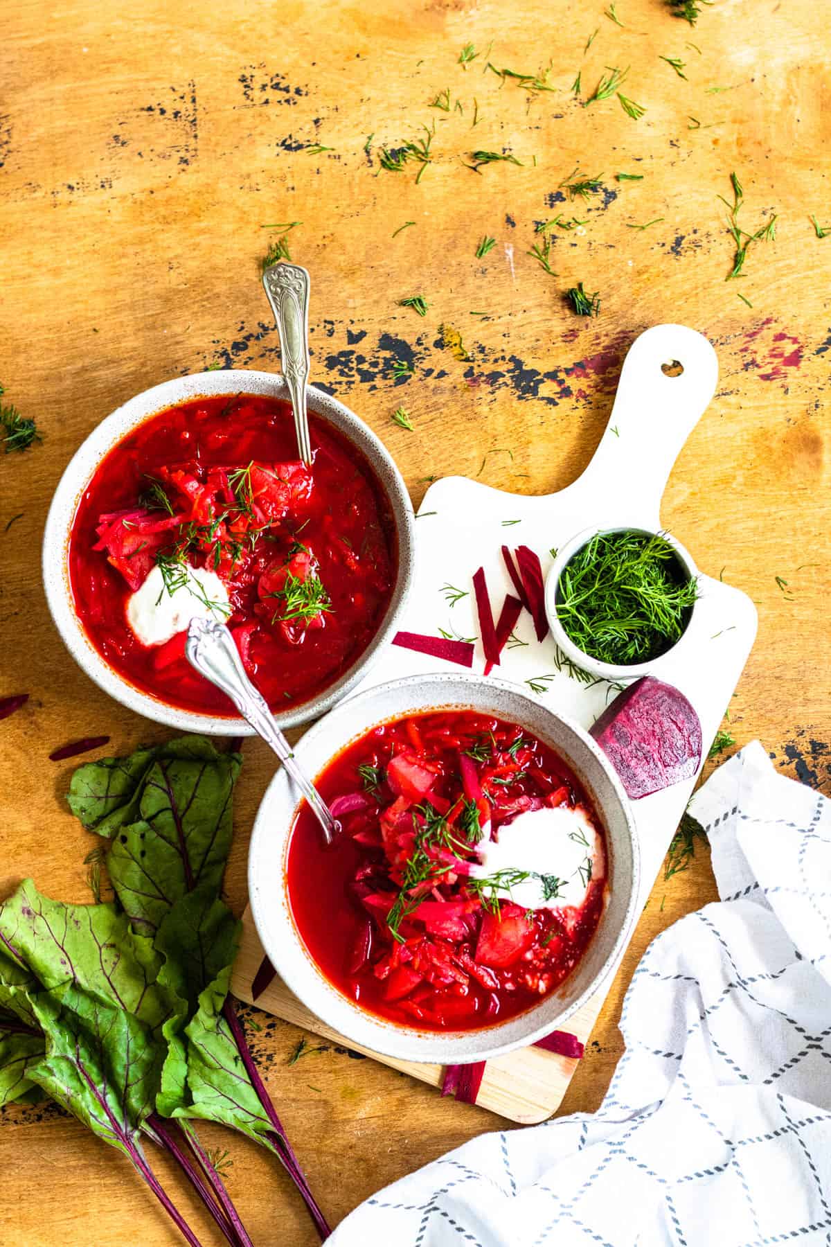 2 bowls of borscht on a cutting board surrounded by dill, a white cloth napkin, beet leaves, and beets. 