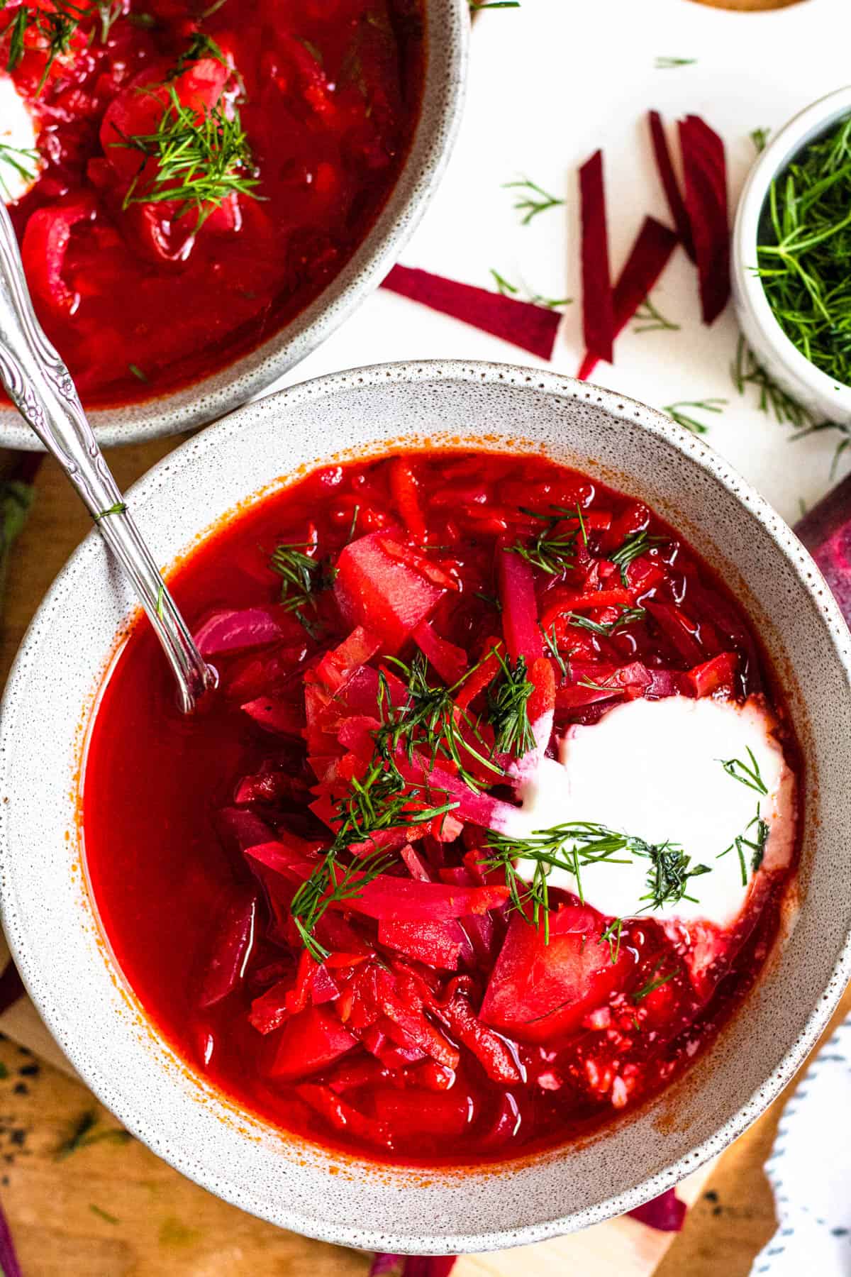 close up of vegetarian borscht soup in a speckled bowl. there is a spoon in the bowl and the soup is topped with dill and sour cream