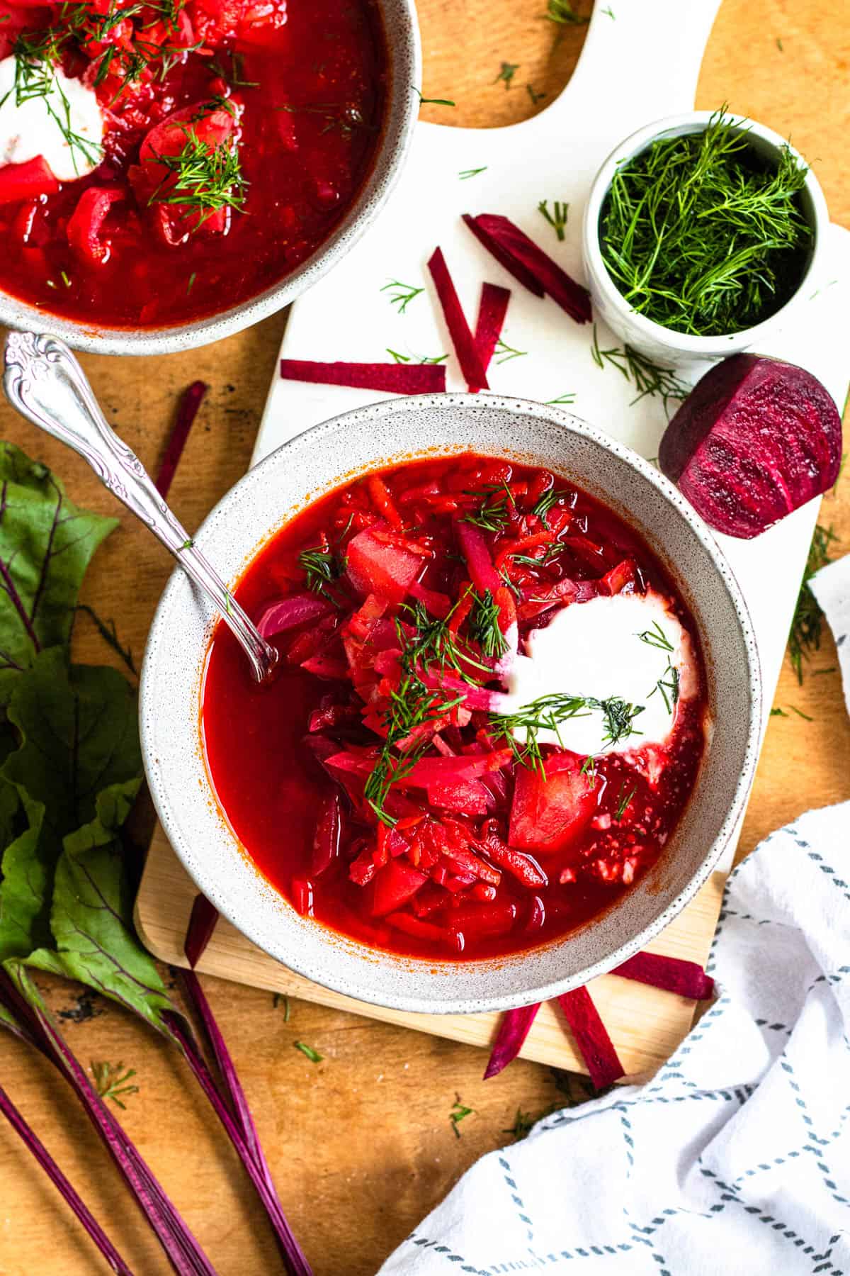 Borsht soup surrounded by dill and fresh beets, displayed on a white cutting board