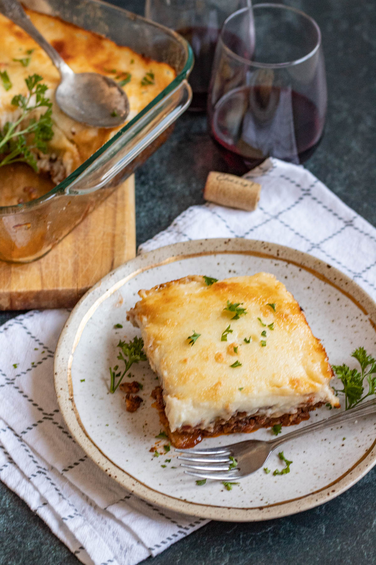 Slice of moussaka sitting on a plate with a fork and parsley. A napkin, wine cork, and moussaka tray are sitting in the background. 