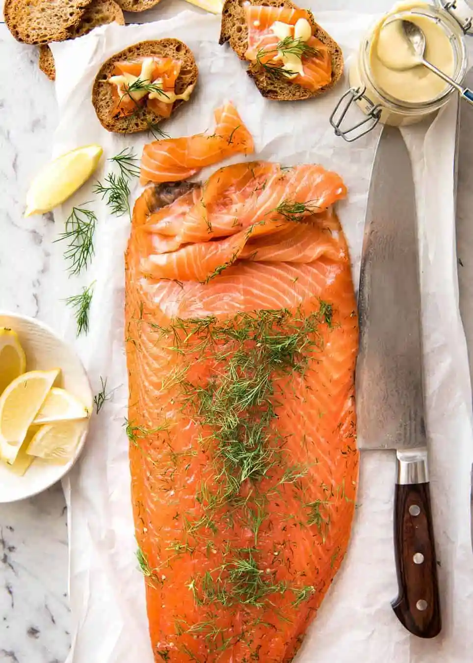 Salmon (gravadlax) served at a Swedish Christmas, on parchment paper next to a chef's knife with fresh dill and crostinis topped with raw salmon and fresh dill with a sauce.  