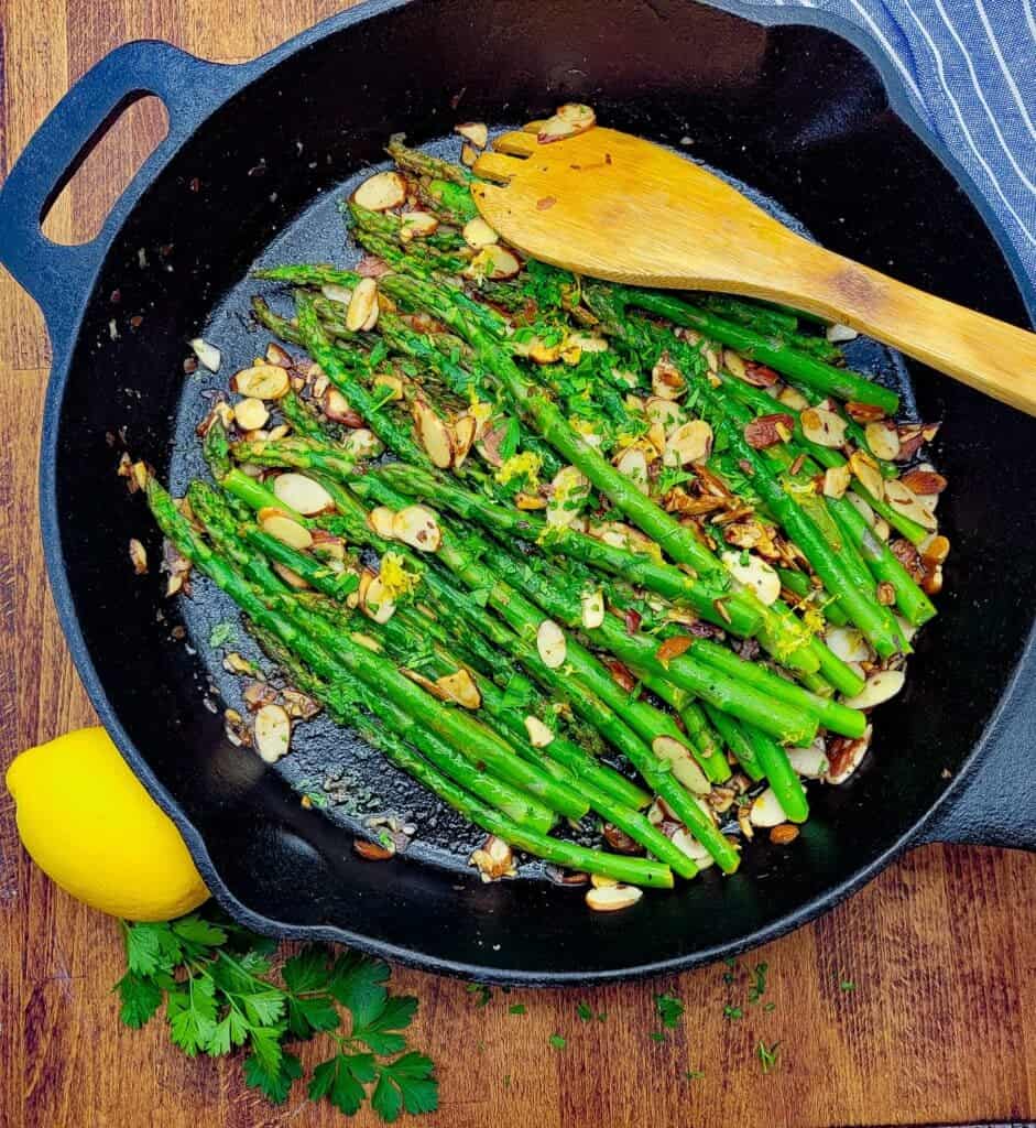 Sauteed asparagus almandine in a cast iron pan with a wooden spoon resting in it. 