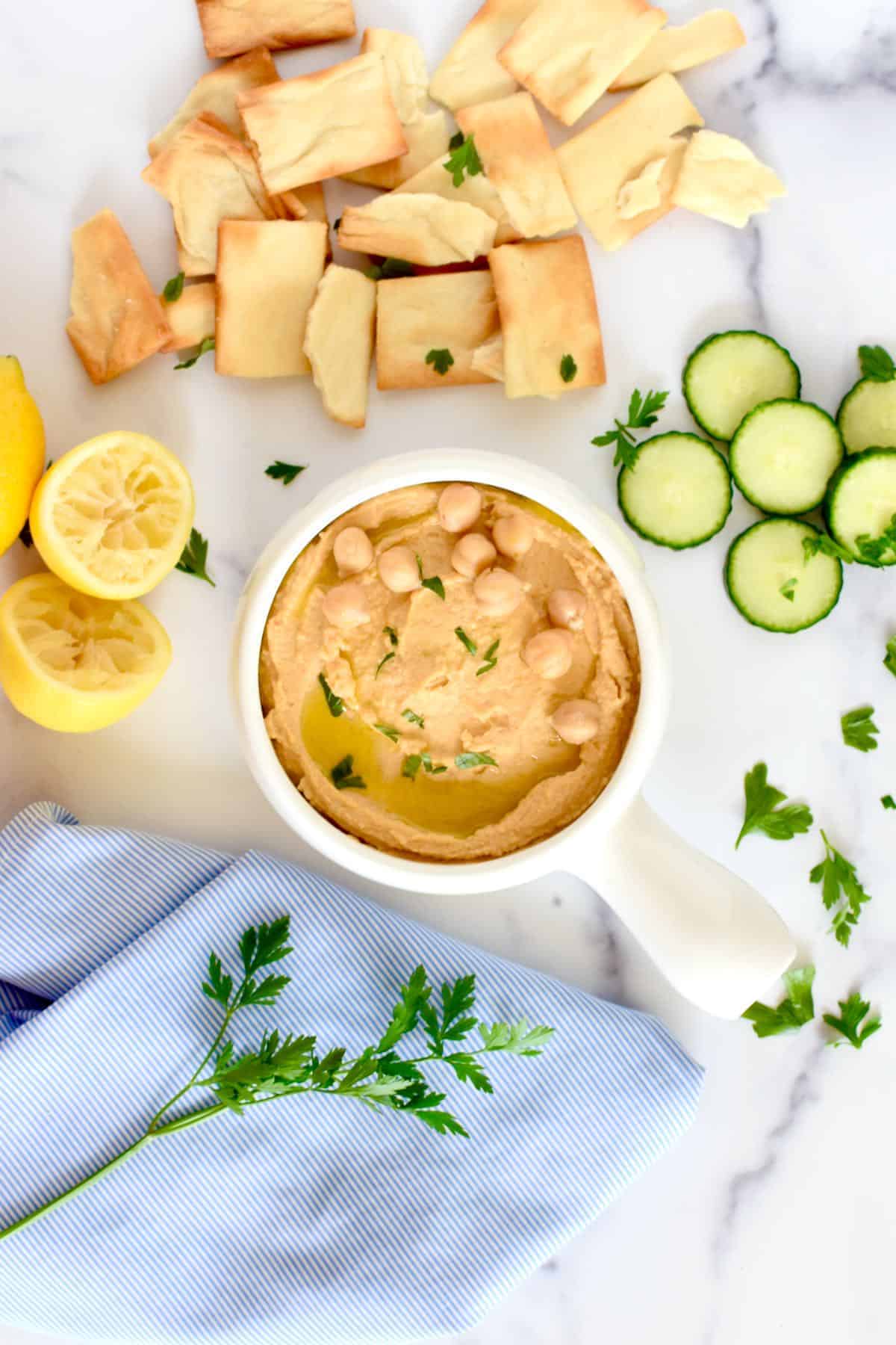 Lemon hummus without garlic in a bowl, garnished with chickpeas and sliced cucumbers and a halved lemon laying next to it. 