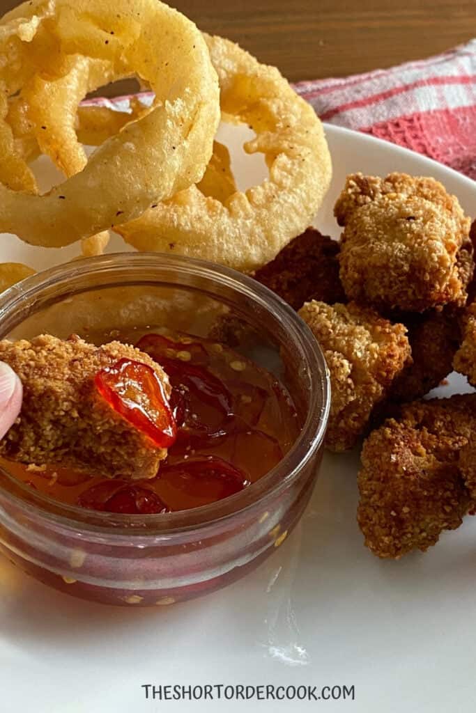 Small bowl of hot honey with chicken nuggets and onion rings on the plat with it. 