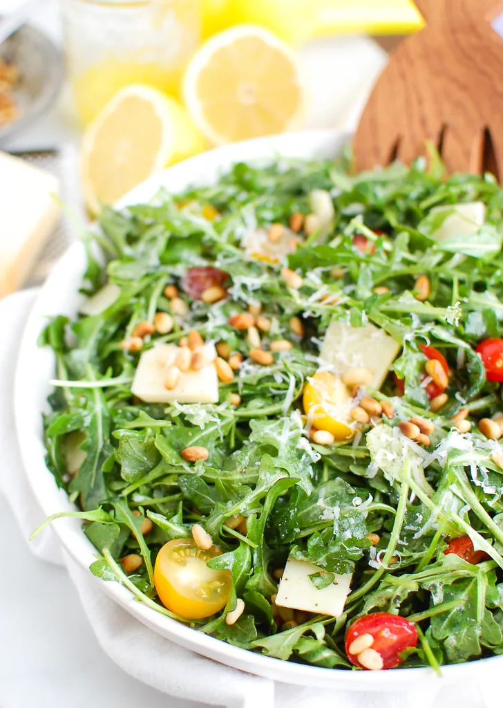 Wooden salad tongs in a bowl of lemon arugula salad with pine nuts. 