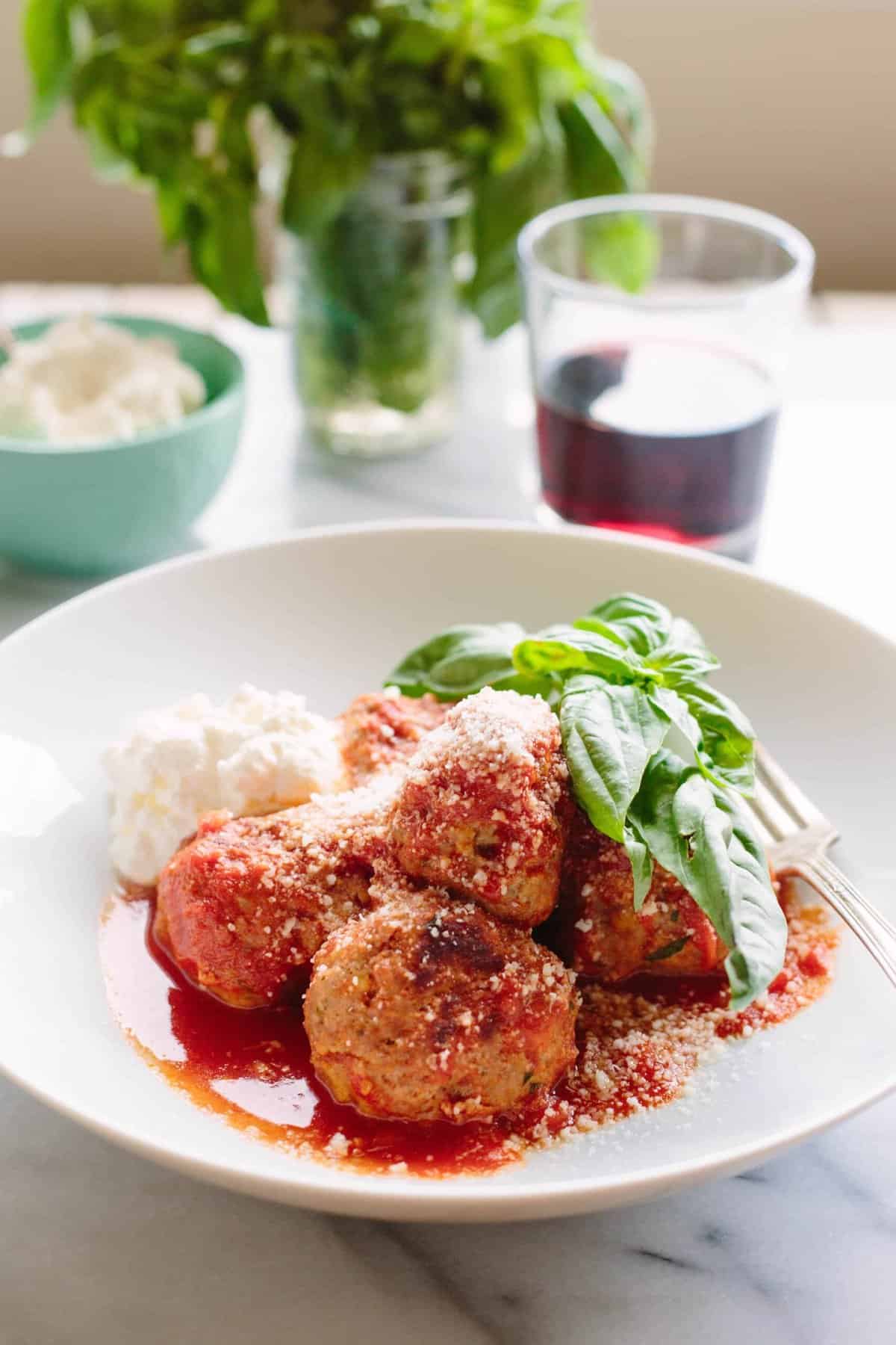 Italian meatballs on a plate with a tomato sauce poured over them with fresh basil leaves and a dollop of ricotta cheese. 