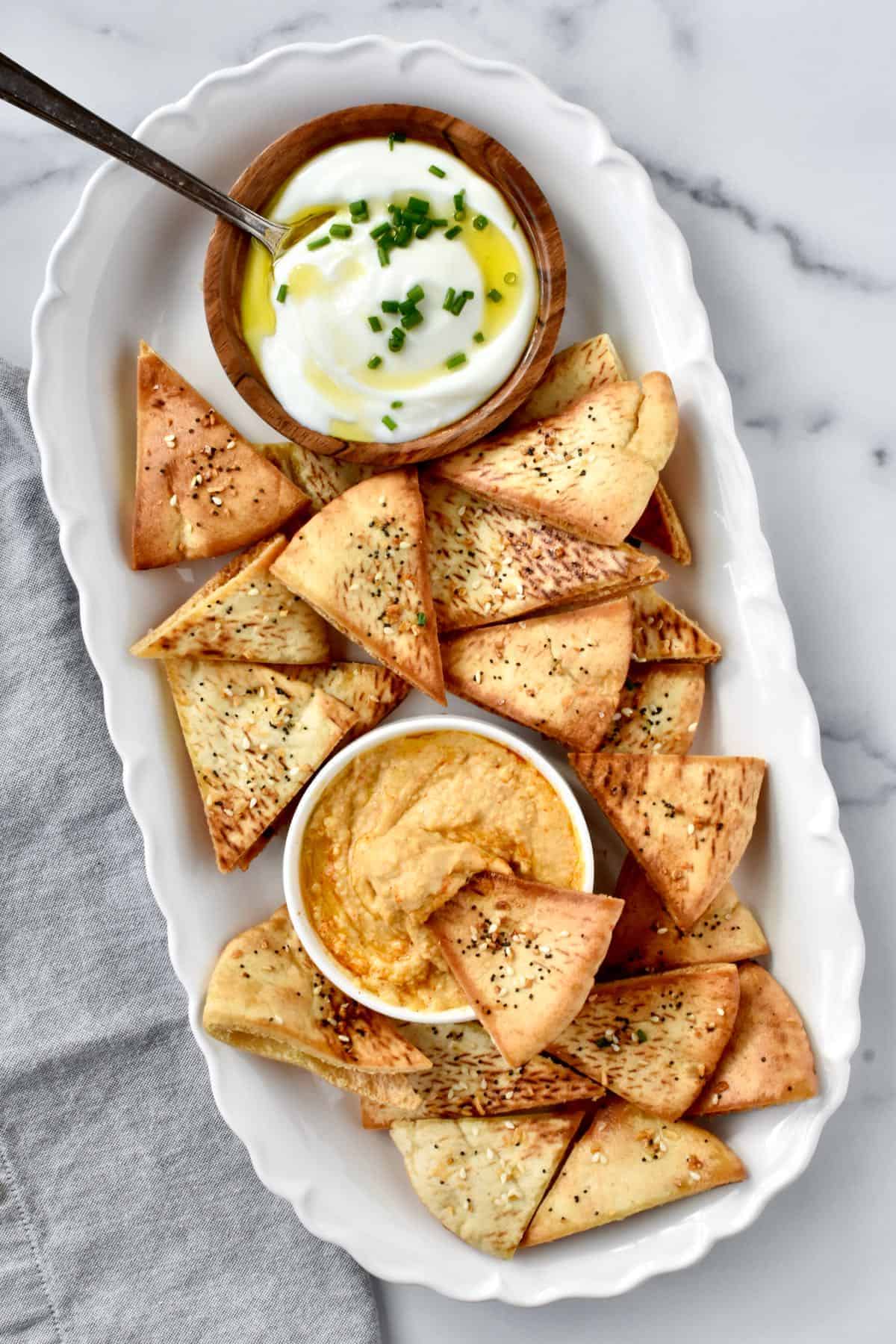 Air fryer pita chips on a platter with a small bowl of hummus and a small bowl of ricotta cheese. 