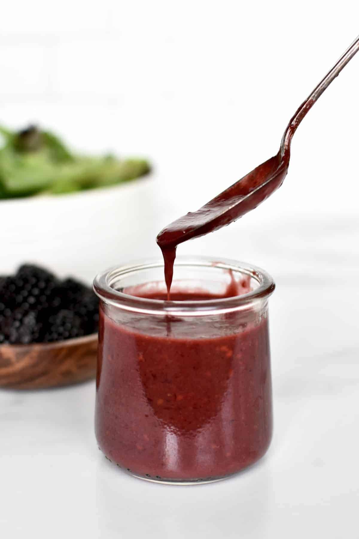 Blackberry balsamic vinaigrette in a small glass jar with a spoonful of it falling off the spoon. 