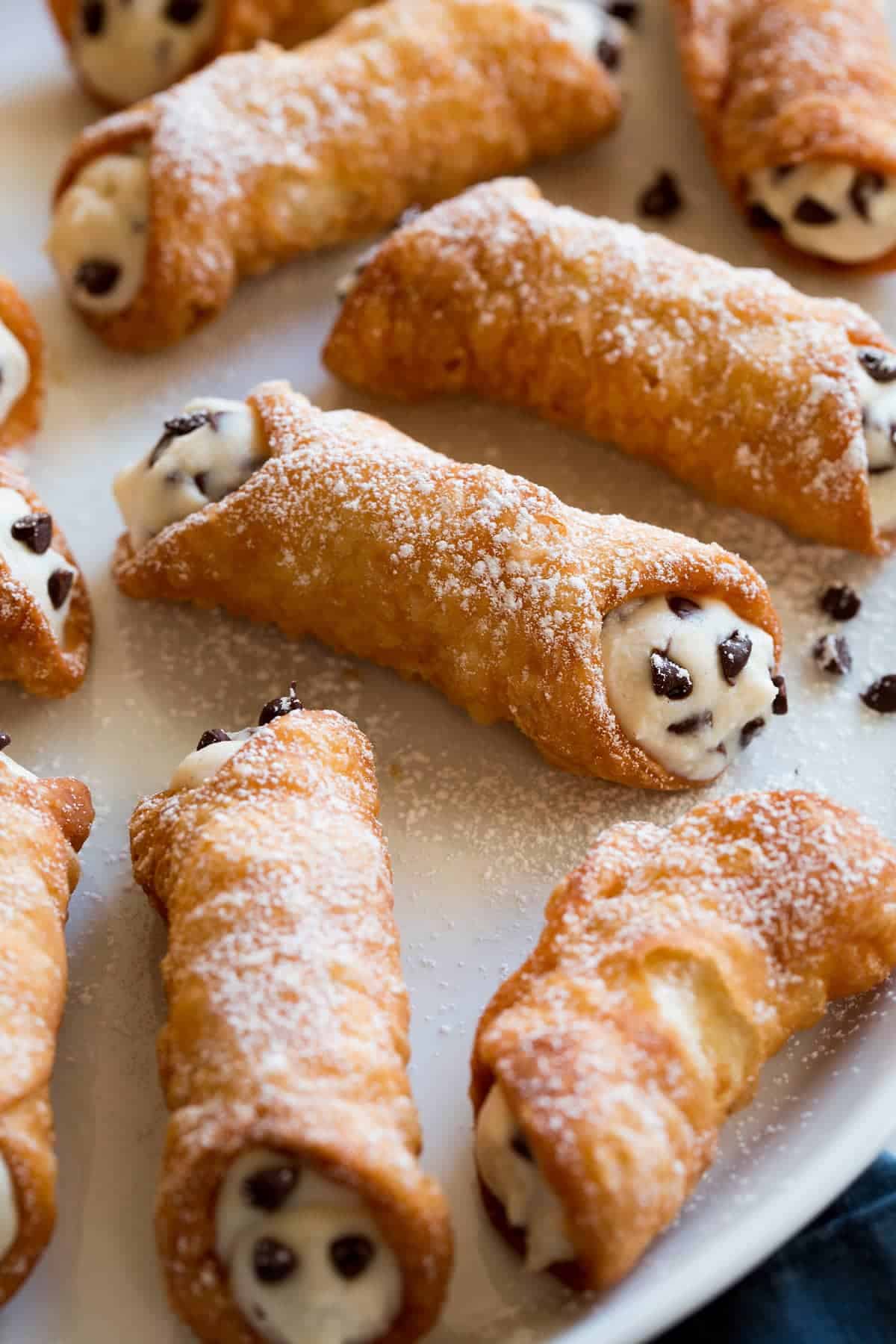Cannolis, an Italian dessert recipe, with mini chocolate chips in crispy shells and powdered sugar sprinkled over the top laying on parchment paper. 