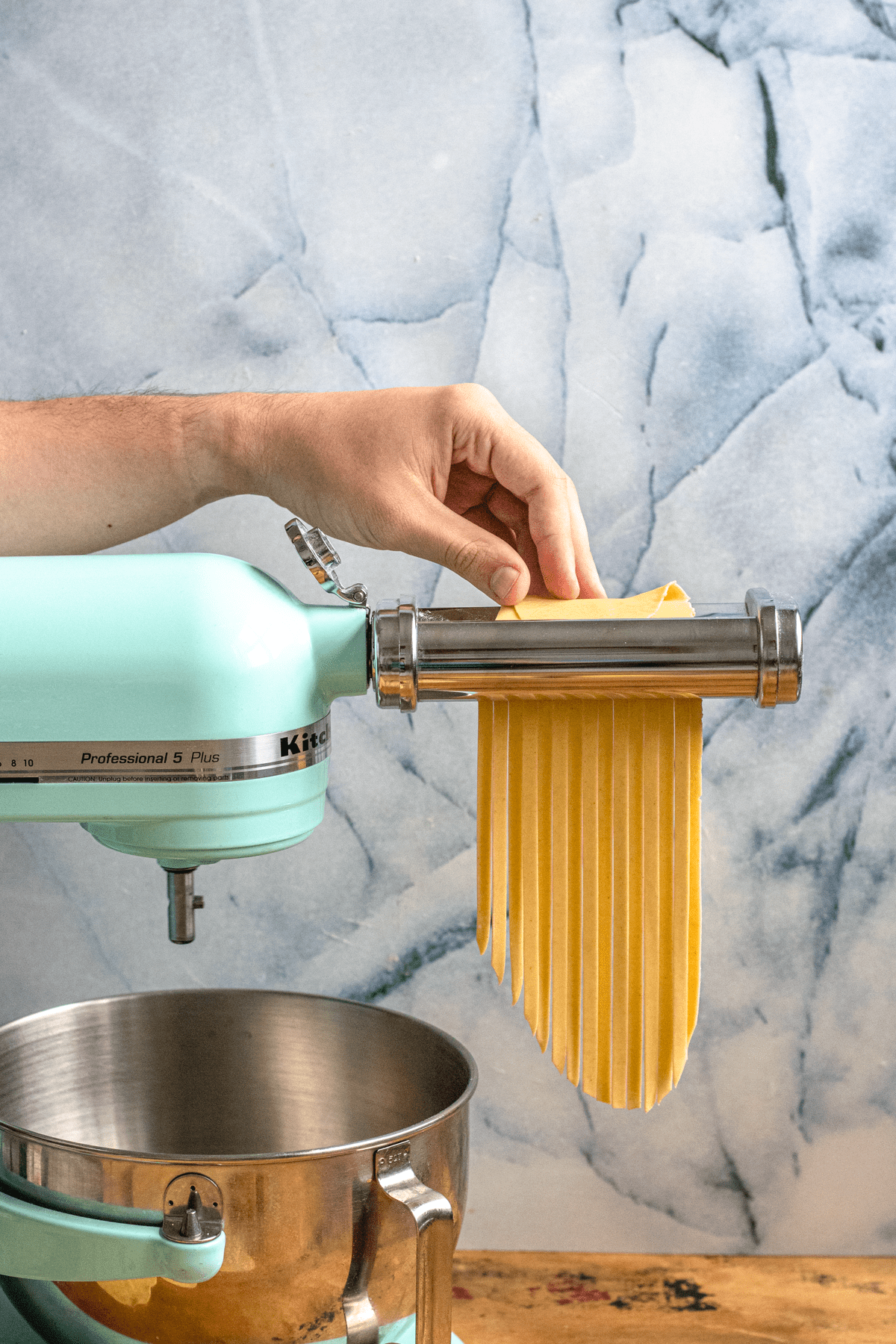A hand running a sheet of pasta  dough sheet through a fettuccine cutter. 