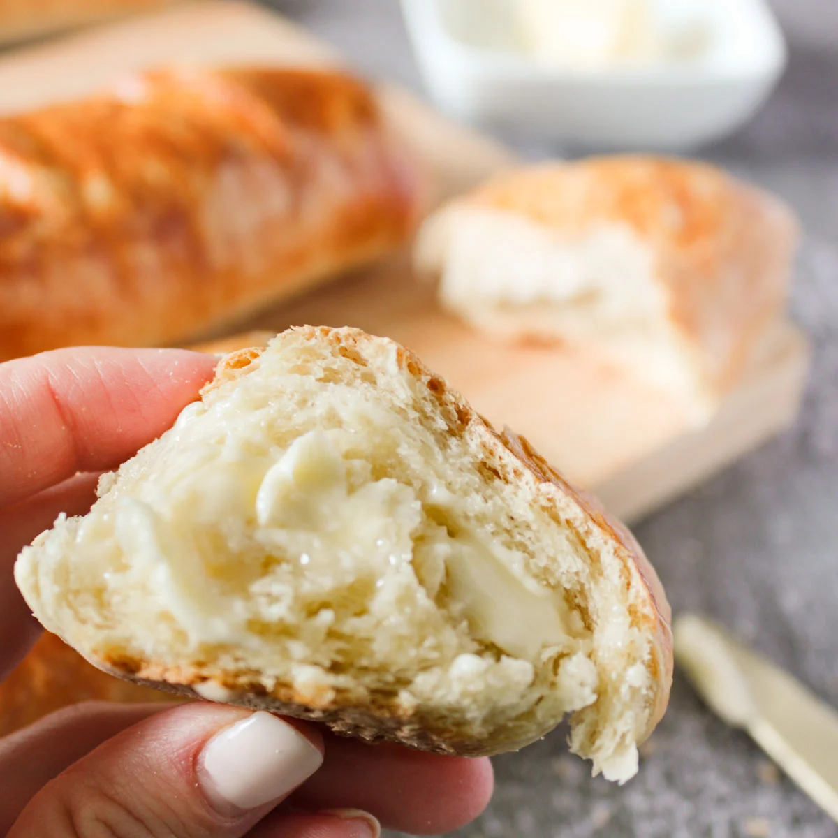 A piece of buttered easy homemade baguette held up by a hand over the loaf. 