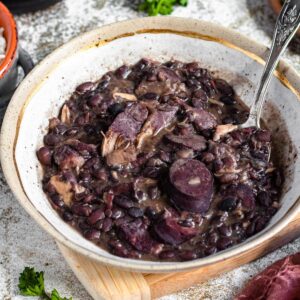 A large bowl of feijoada in a bowl on a cutting board with a spoon resting in the stew.